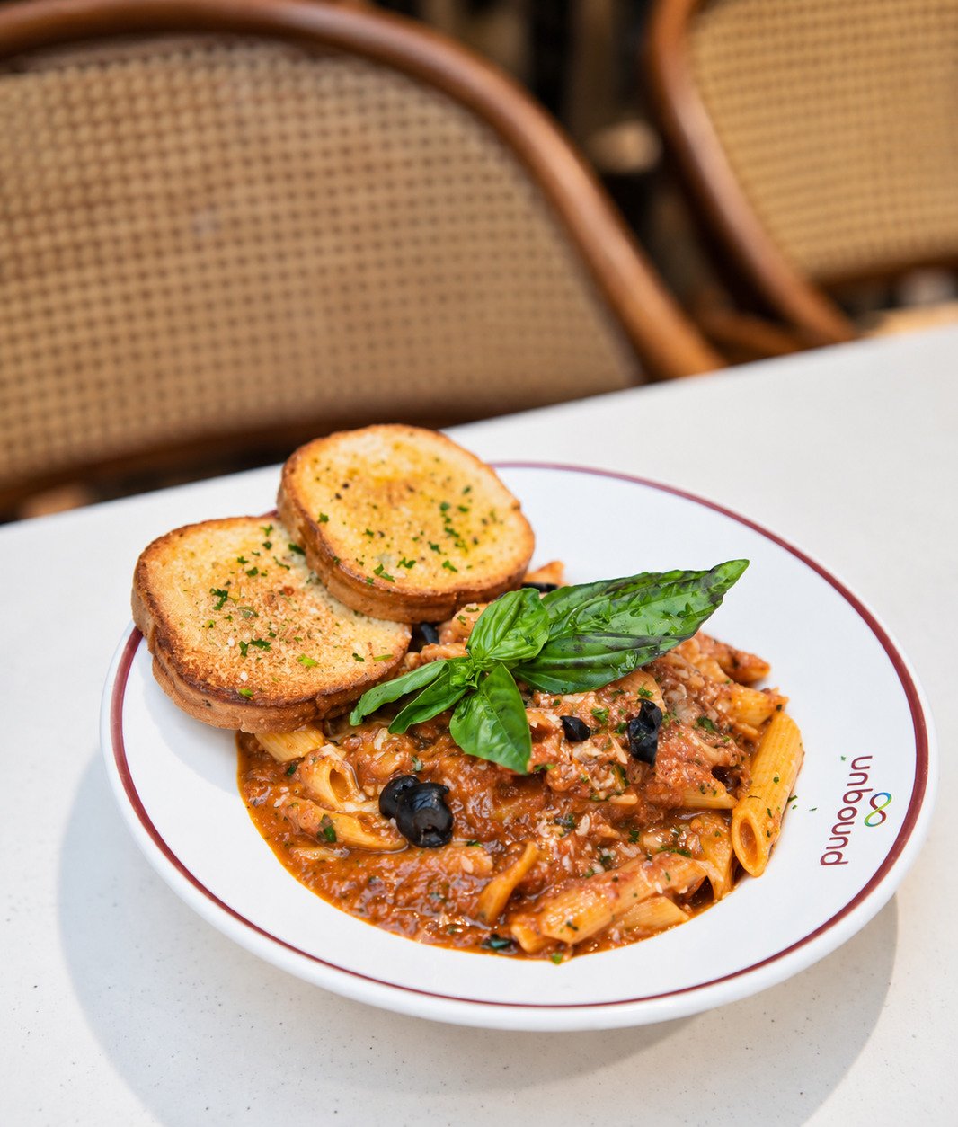 A bowl of penne in a red sauce with basil, olives and two slices of garlic bread
